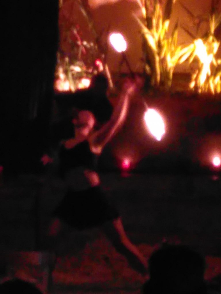 Das Bild ist verschwommen und von Dunkelheit und warmen Farben geprägt. Im Vordergrund ist eine Person zu sehen, die mit Feuer jongliert, im Hintergrund beleuchtetes Stroh, der Farbverlauf ist von links und unten von schwarz über rot zu orange.